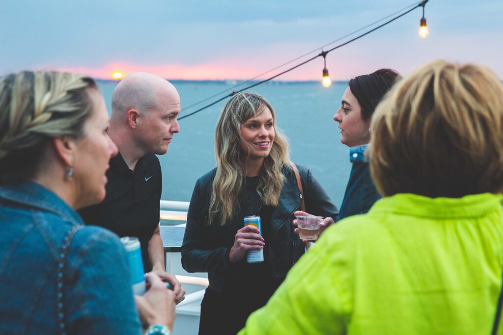 Group of five people holding drinkis and talking with water and sunset in the background.